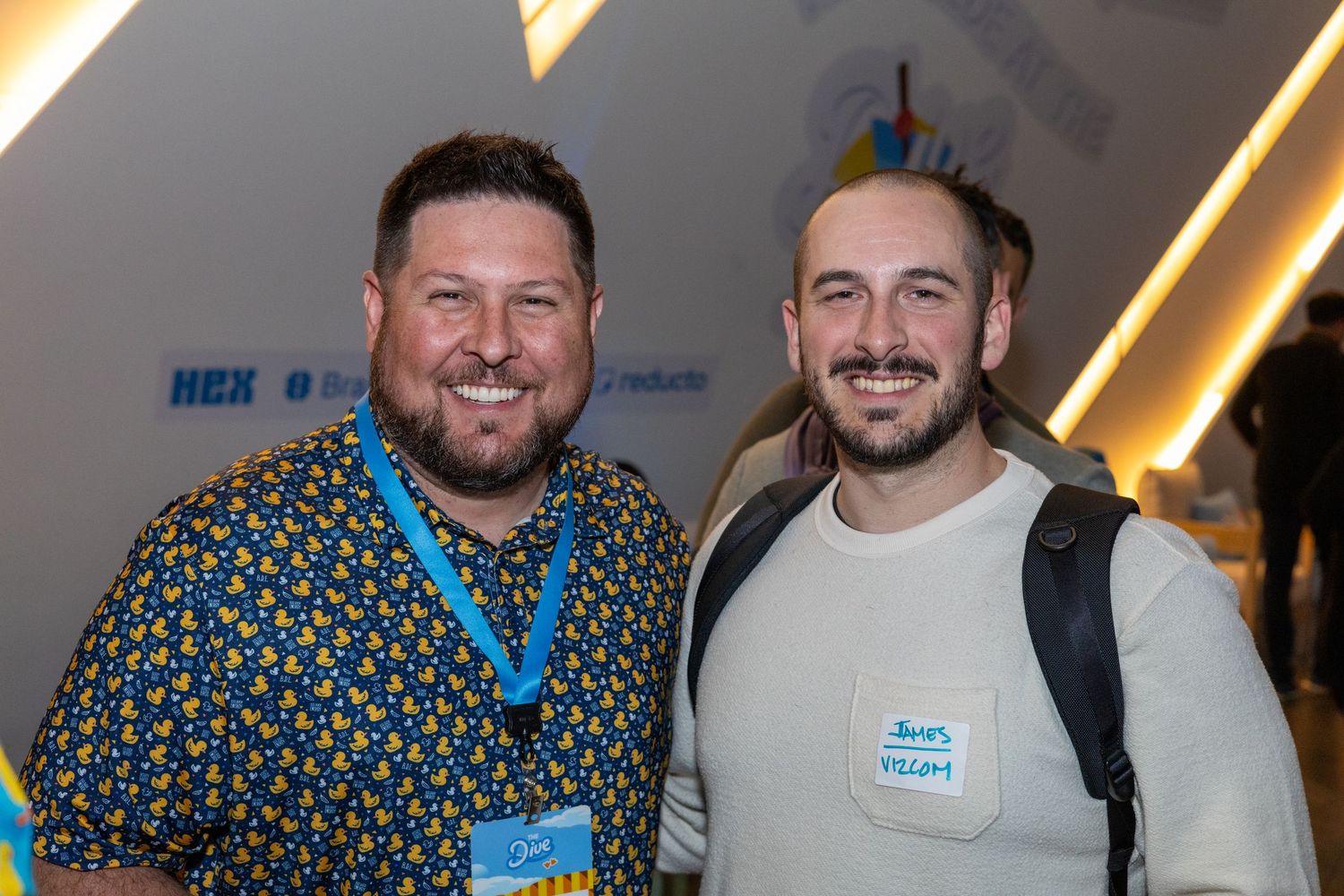 Two smiling men wearing name tags pose together at a professional networking event with colorful backdrop.