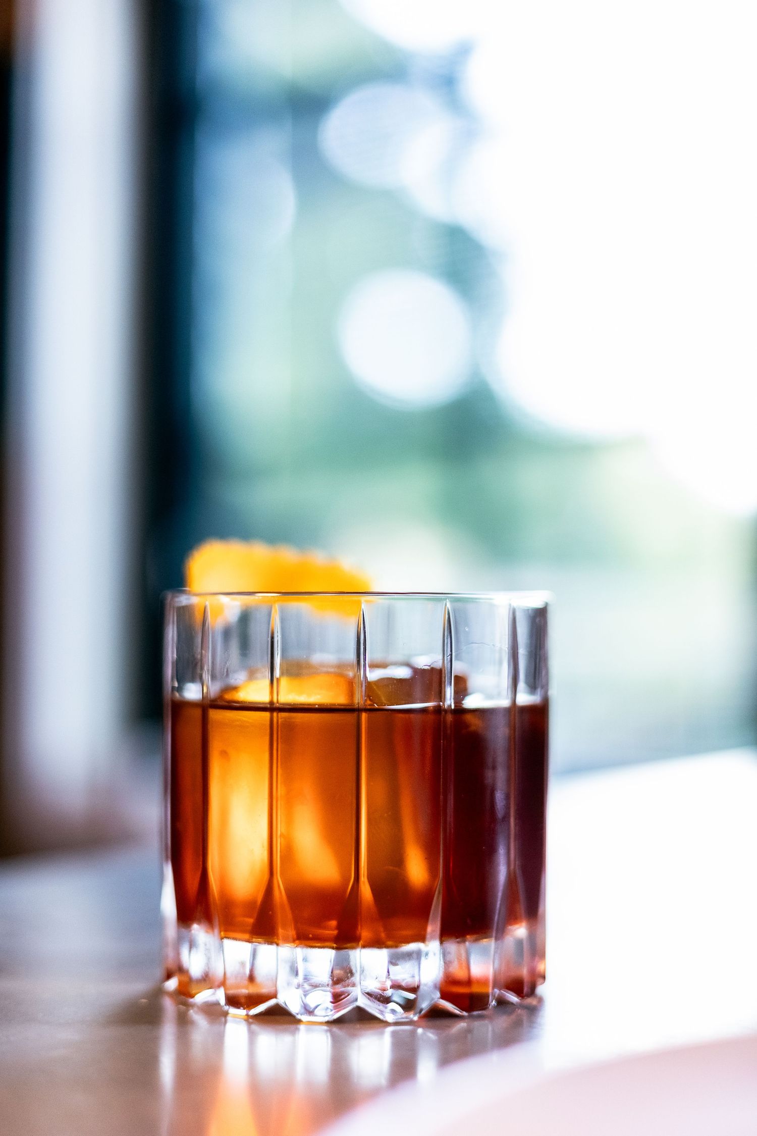 Classic Old Fashioned cocktail with orange peel garnish in a crystal glass by a sunny window.