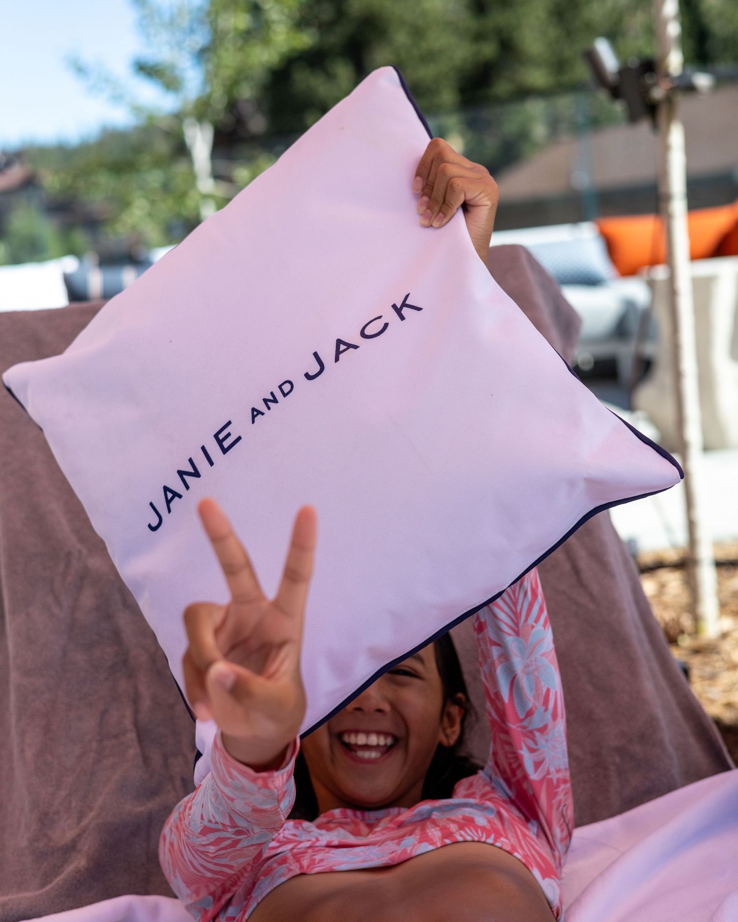 Smiling girl holding a white Janie and Jack branded pillow while making a peace sign outdoors.