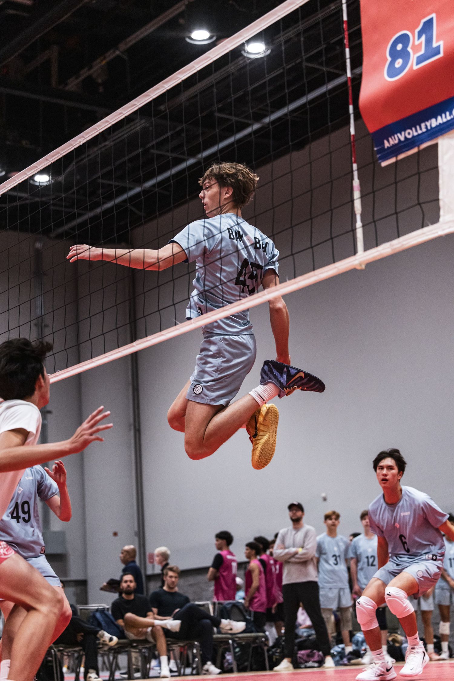 Male volleyball player leaping high at the net during an indoor tournament match, teammates ready in the background.