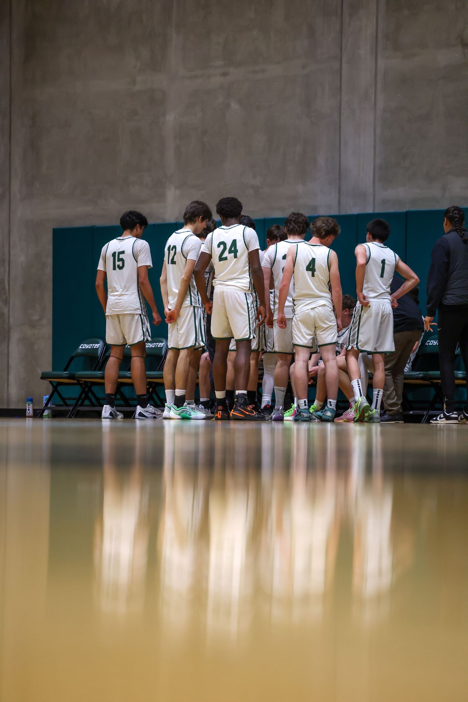 Basketball team huddles on sideline bench in white and green uniforms during a game on a shiny hardwood court.