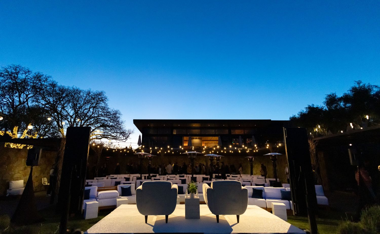 Elegant outdoor evening venue with string lights, white seating, and a glowing pavilion under a twilight blue sky.