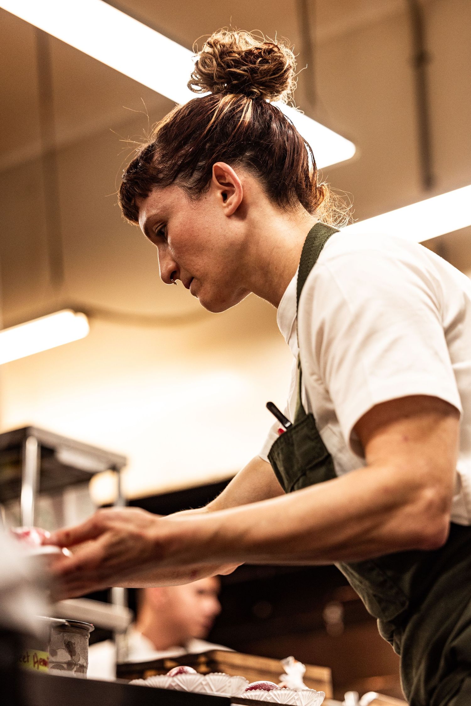 Female chef in green apron and white shirt carefully plating food in a professional kitchen.