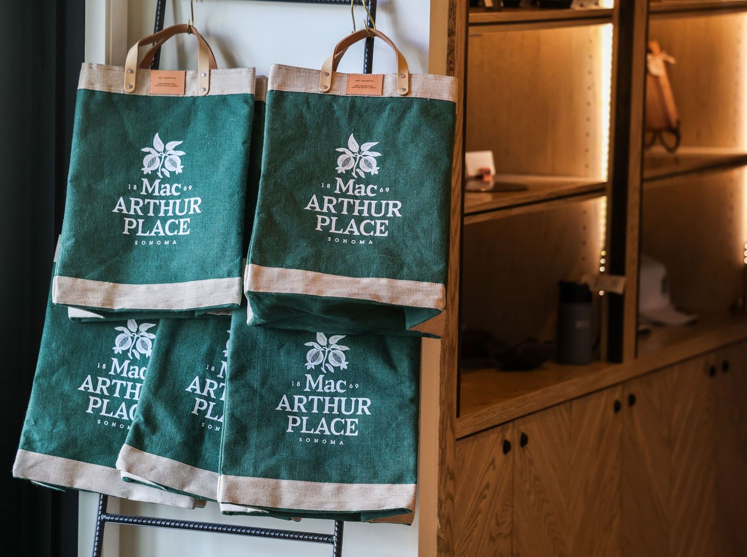 Green MacArthur Place Sonoma branded canvas tote bags hanging on a wooden shelf.