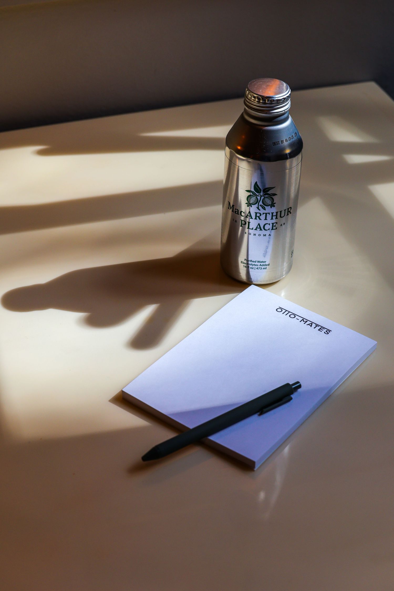 MacArthur Place water bottle beside an Ottorates notepad and pen on a beige desk.