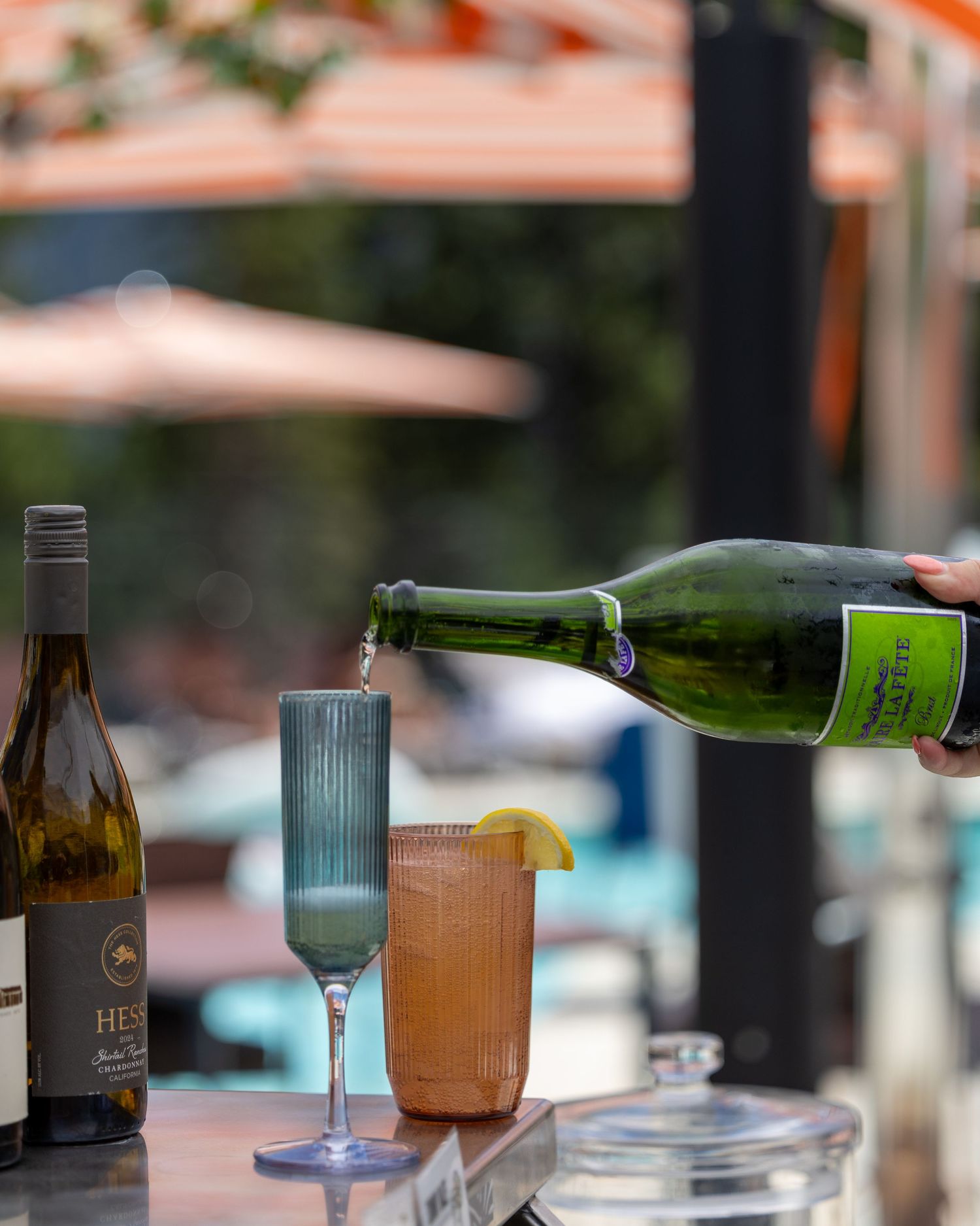 Sparkling wine being poured into a cocktail glass beside a wine bottle at an outdoor poolside bar.