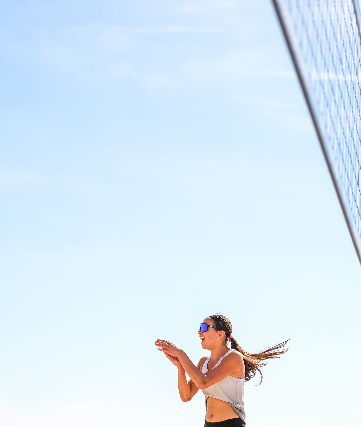 Woman in sunglasses playing beach volleyball, bumping ball near net under clear blue sky.