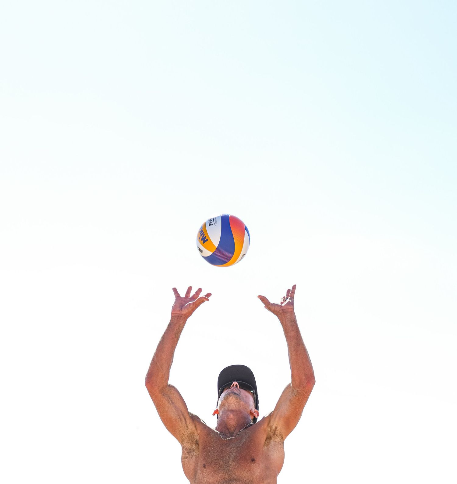 Shirtless man wearing a cap sets a volleyball overhead on a sunny beach day.