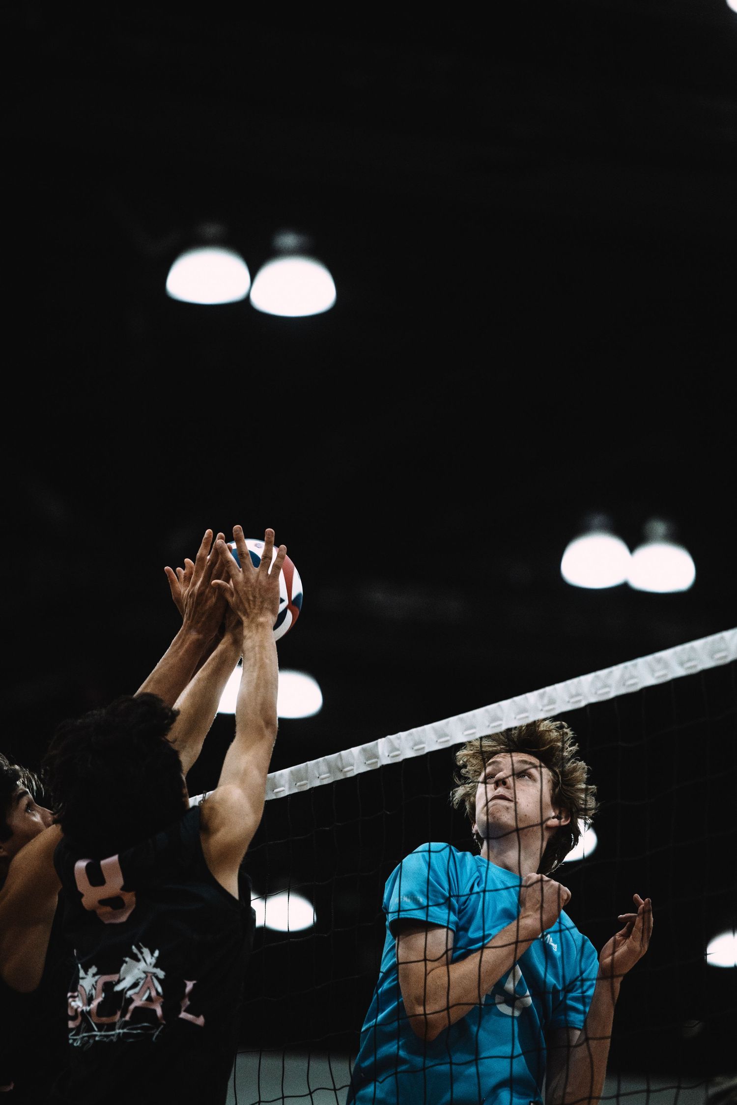Players compete at a volleyball net under bright indoor lights, with one player in a blue shirt watching the block.