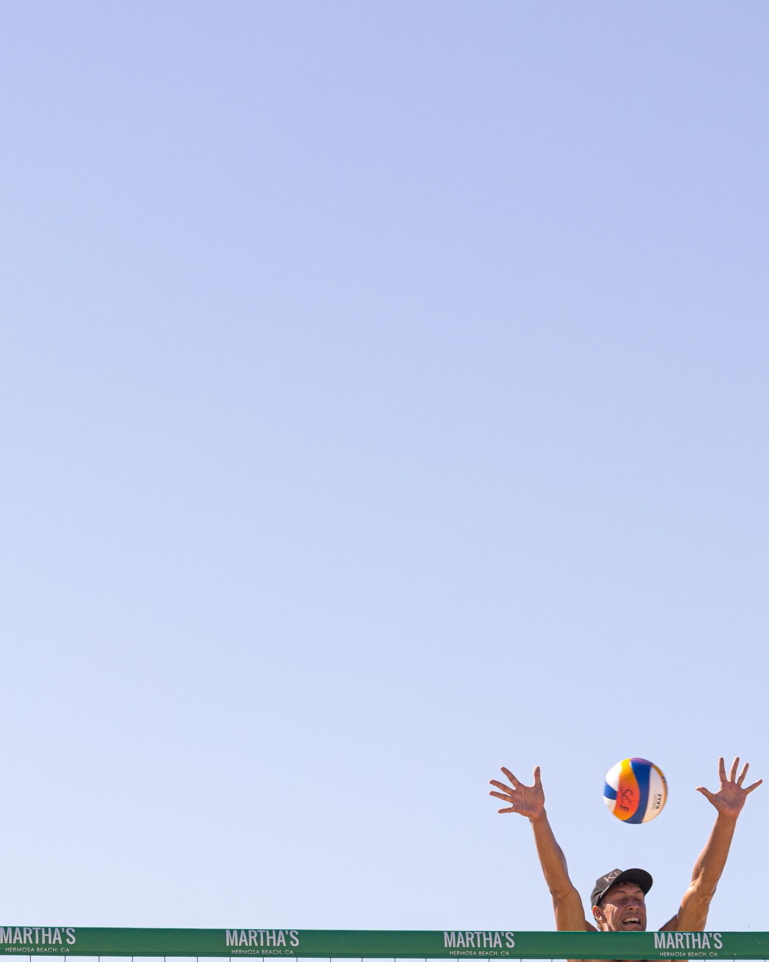 Beach volleyball player jumps with arms raised to set a colorful ball against a clear blue sky at Martha's tournament.