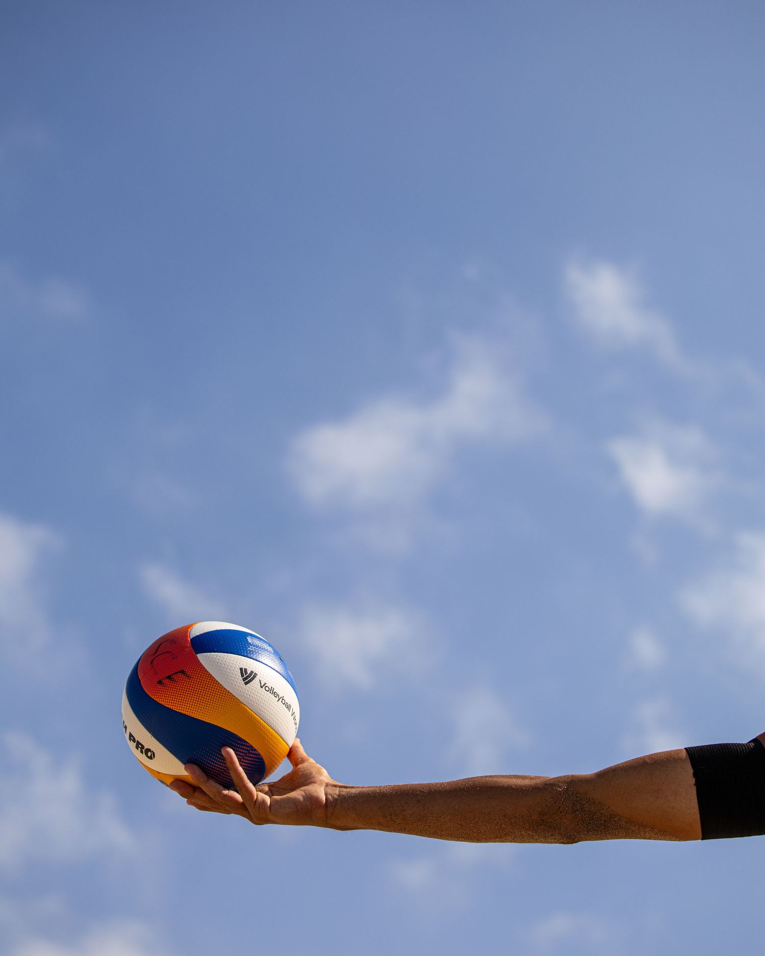 A hand holding a colorful FIVB volleyball up against a partly cloudy blue sky.