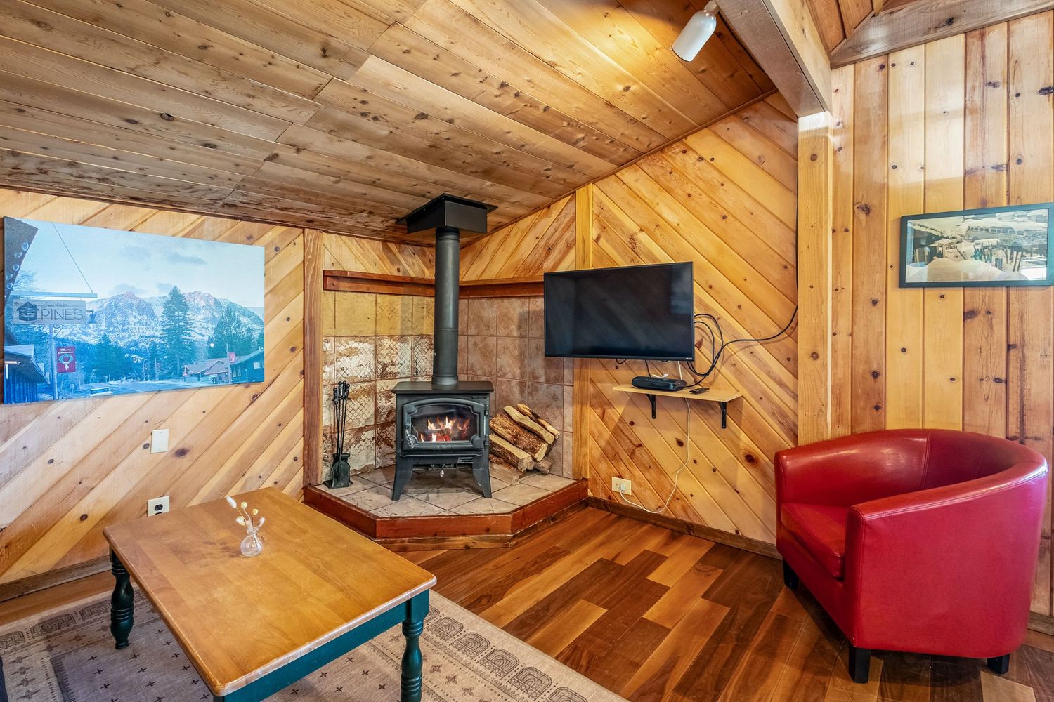 Rustic wood-paneled cabin living room with a black wood-burning stove, TV, red armchair, and hardwood floors.