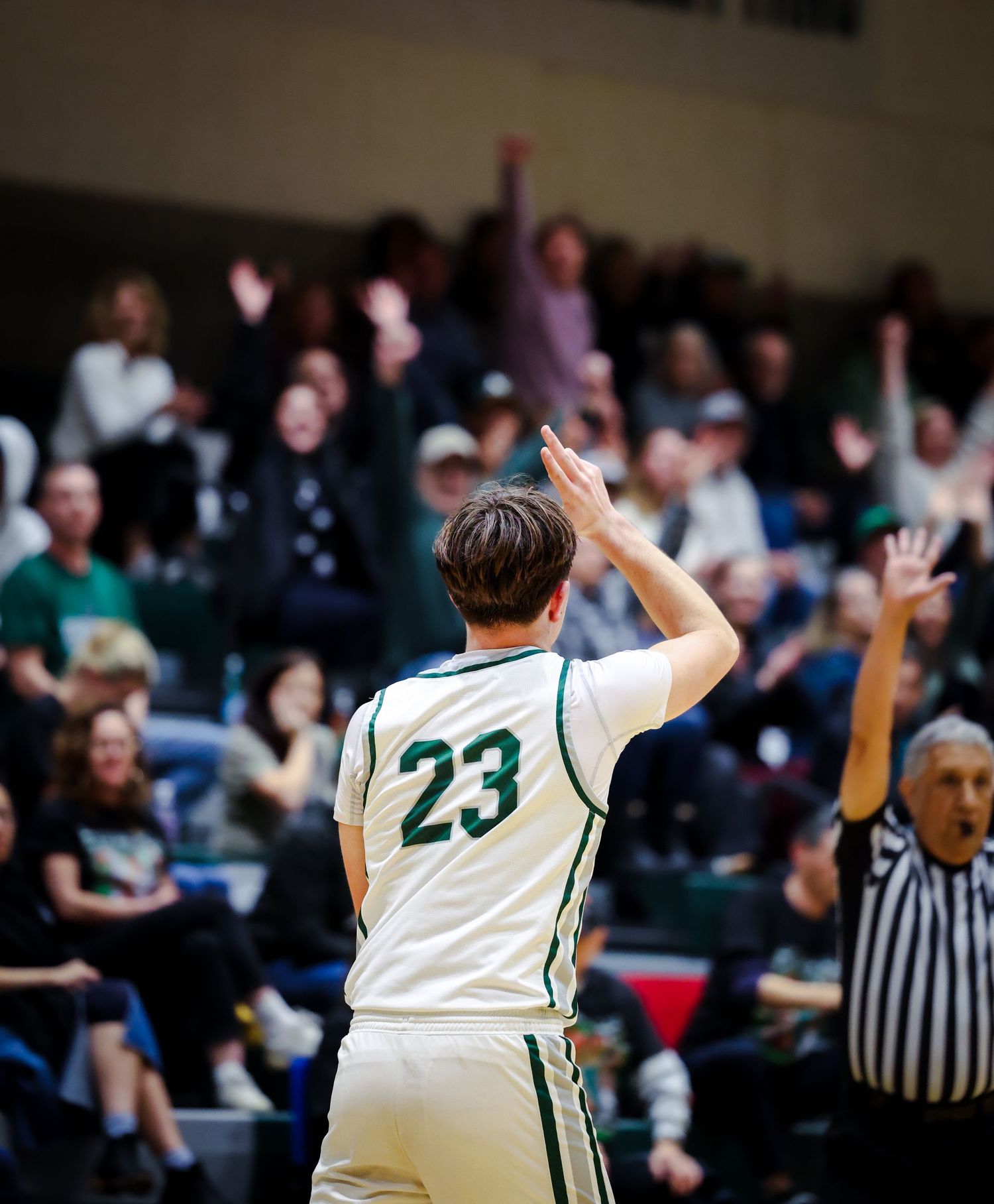 Basketball player #23 raises hands celebrating with excited crowd and referee visible in background.