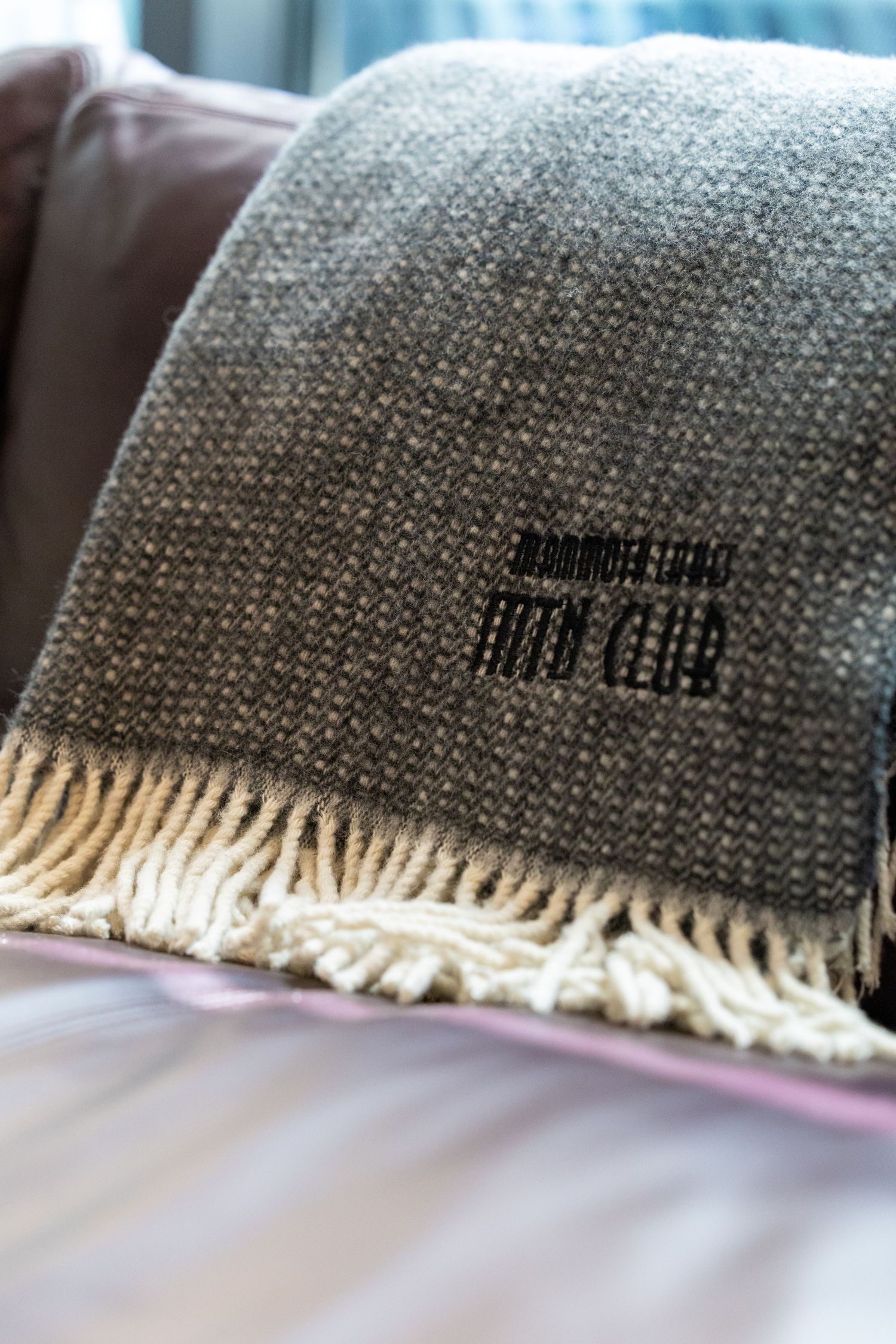 Gray woven throw blanket with fringe and embroidered logo draped over a pink sofa.