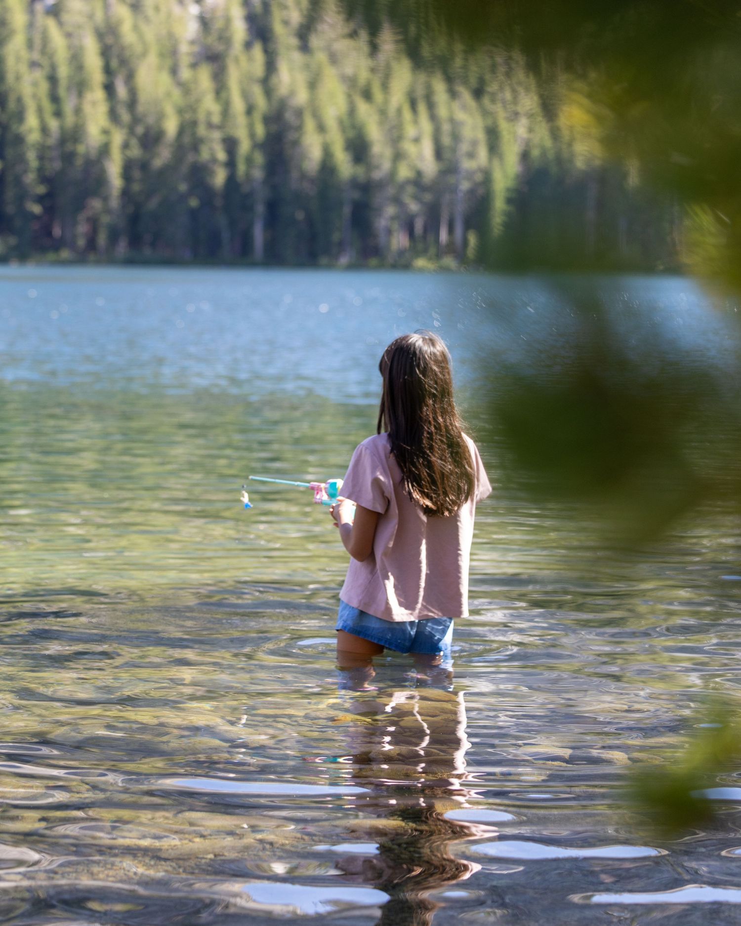 A young girl wades in a serene mountain lake surrounded by pine trees.