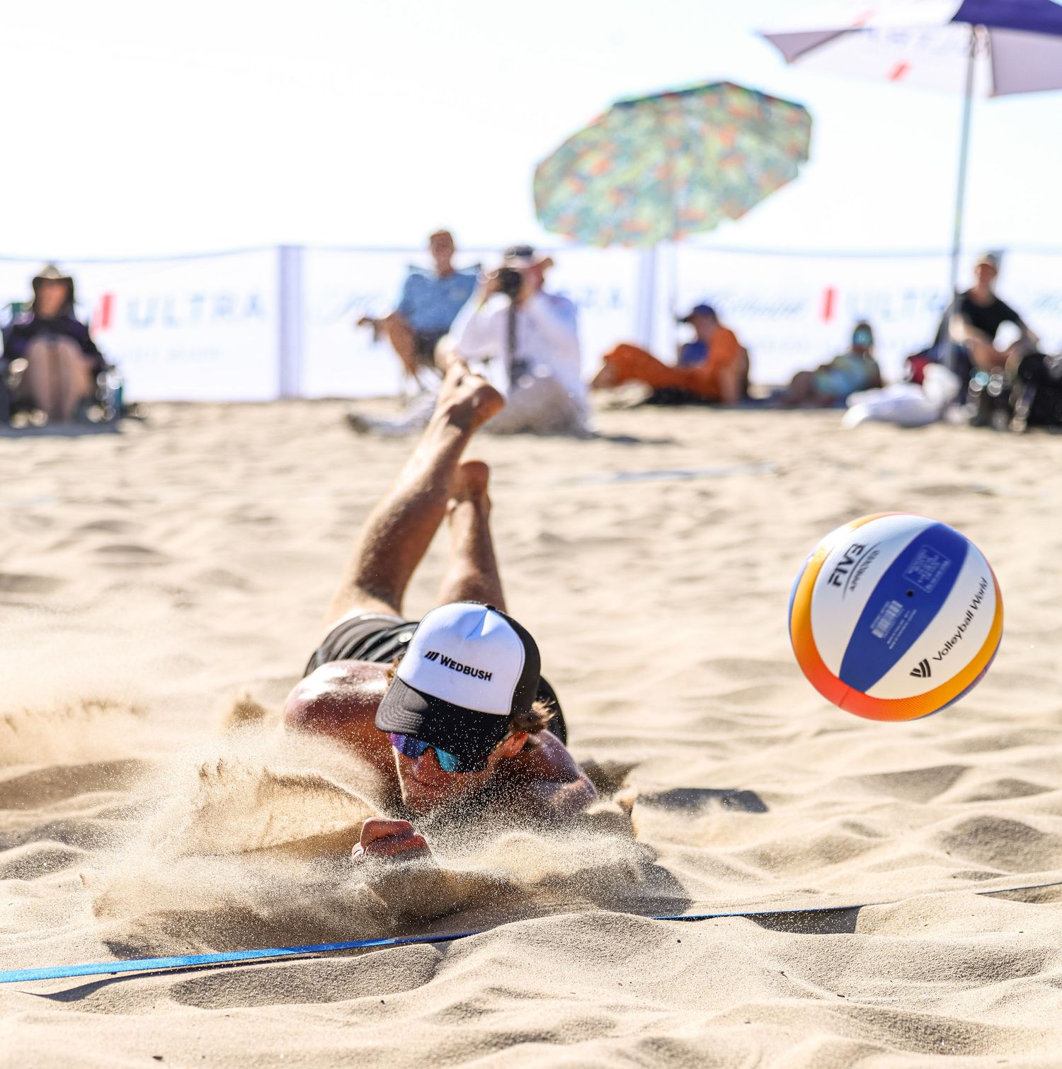 Beach volleyball player diving into sand to save ball during outdoor tournament match.