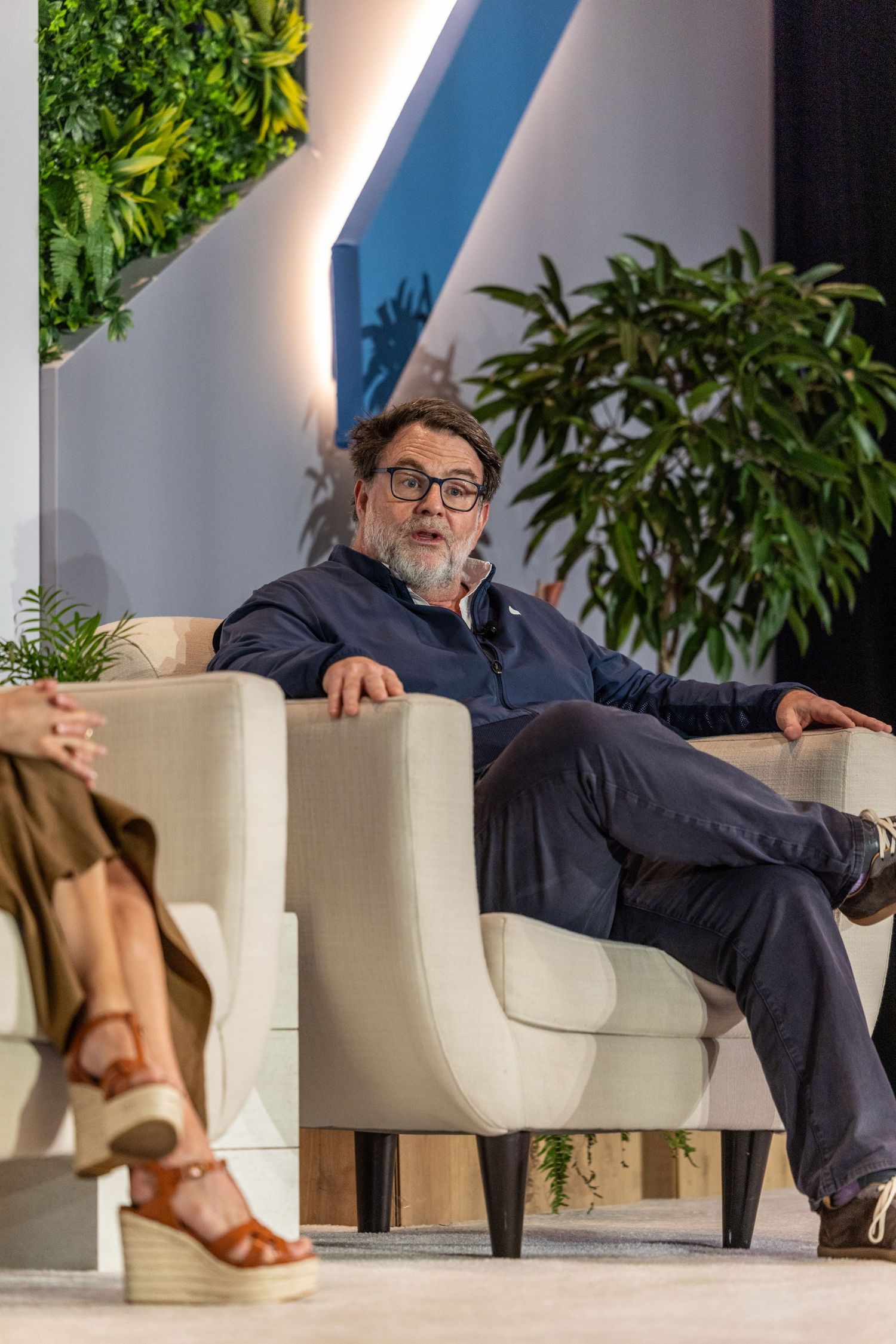 Man with glasses sits relaxed in white armchair during panel discussion, green plant wall backdrop at conference event.