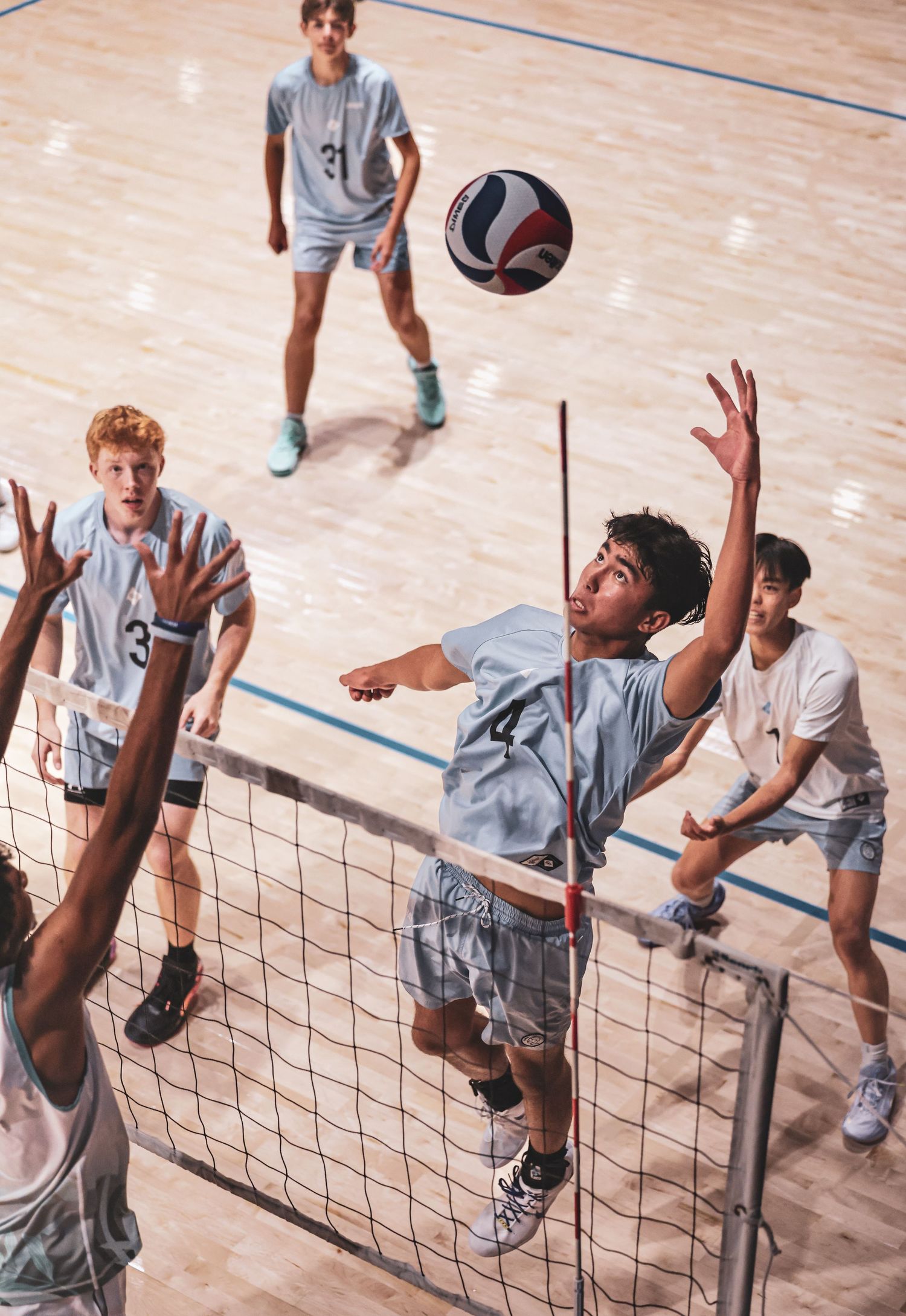 Young men playing competitive indoor volleyball, with one player jumping to spike the ball over the net during a match.