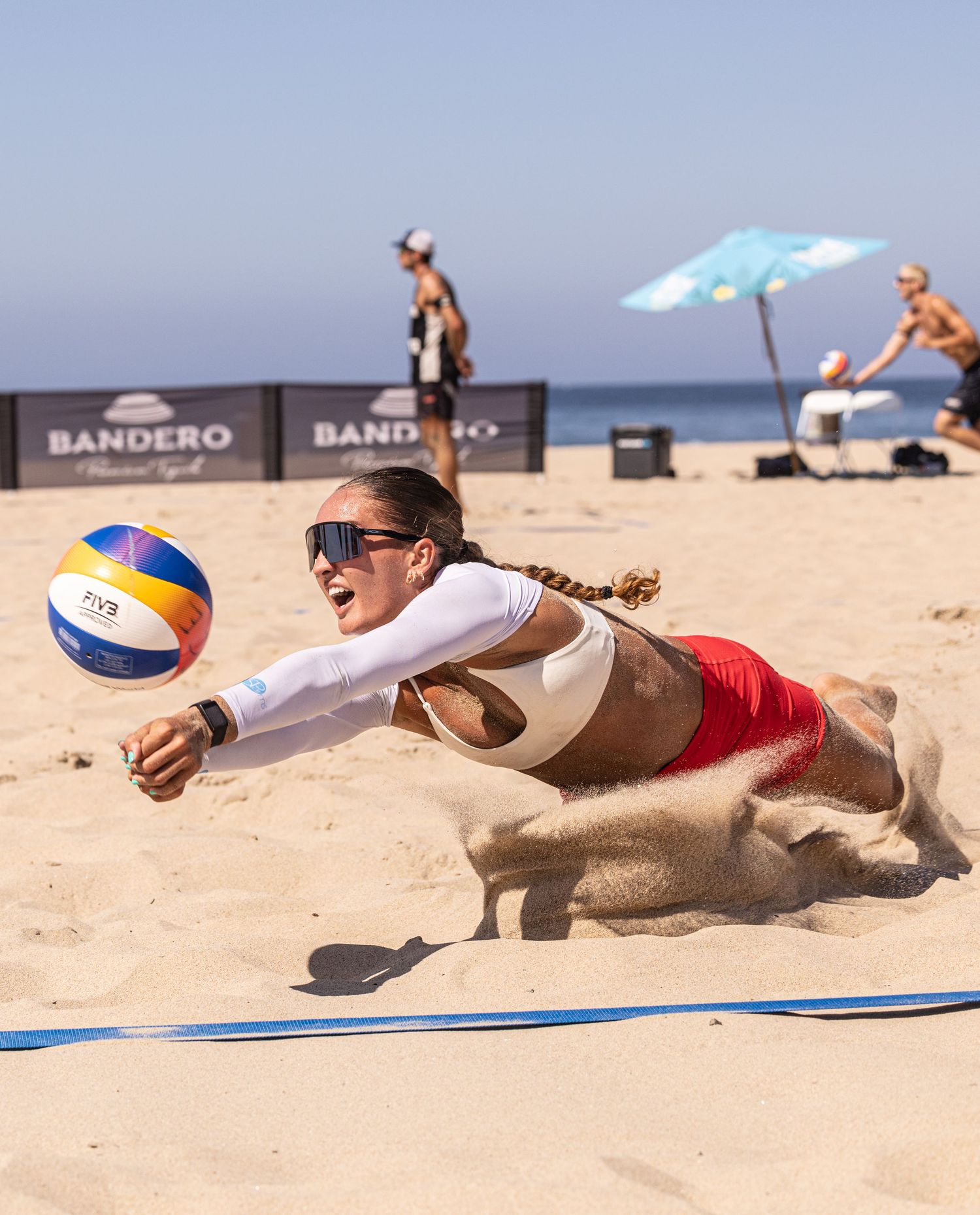 Female beach volleyball player diving into sand to dig a ball during a competitive match with Bandero sponsorship banners.