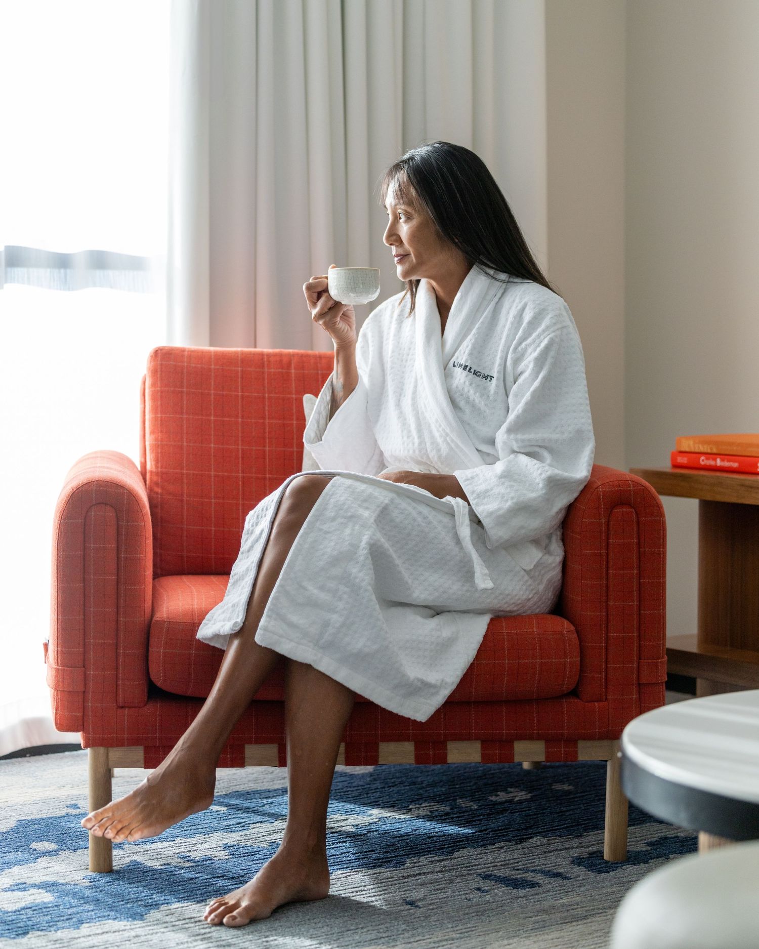Woman in white robe sitting in red chair drinking coffee.