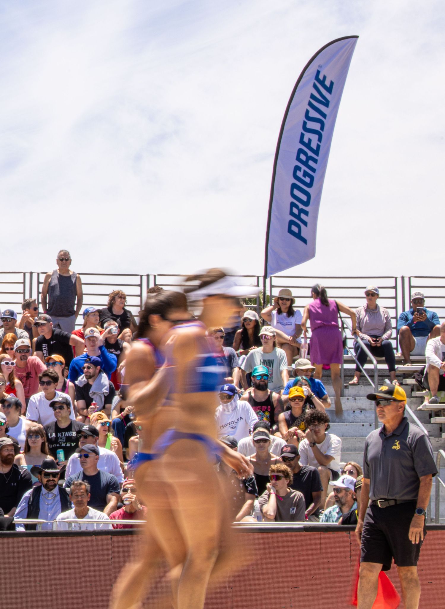 Beach volleyball players compete at a Progressive-sponsored tournament with a packed crowd of spectators in the stands.