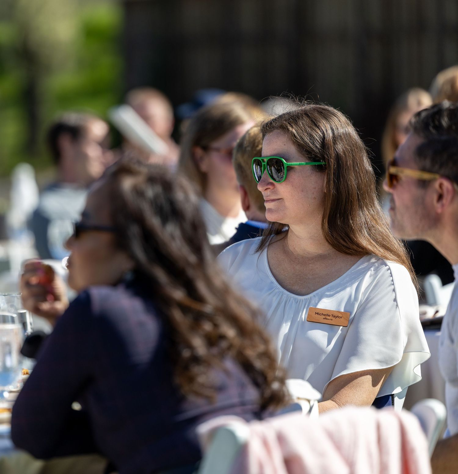 Woman wearing green sunglasses and white top sits at outdoor dining event with blurred guests in background.