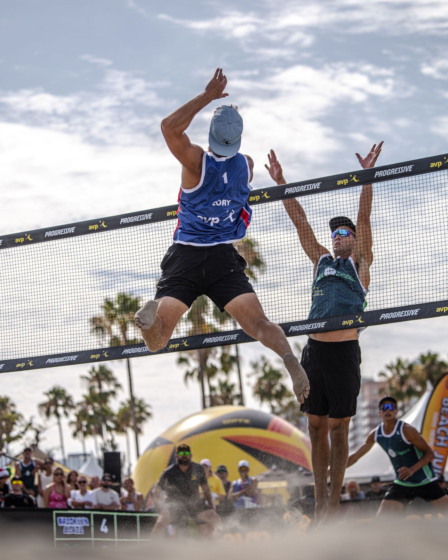 Two men compete in a beach volleyball match, one jumping to spike the ball over the net at an outdoor tournament.