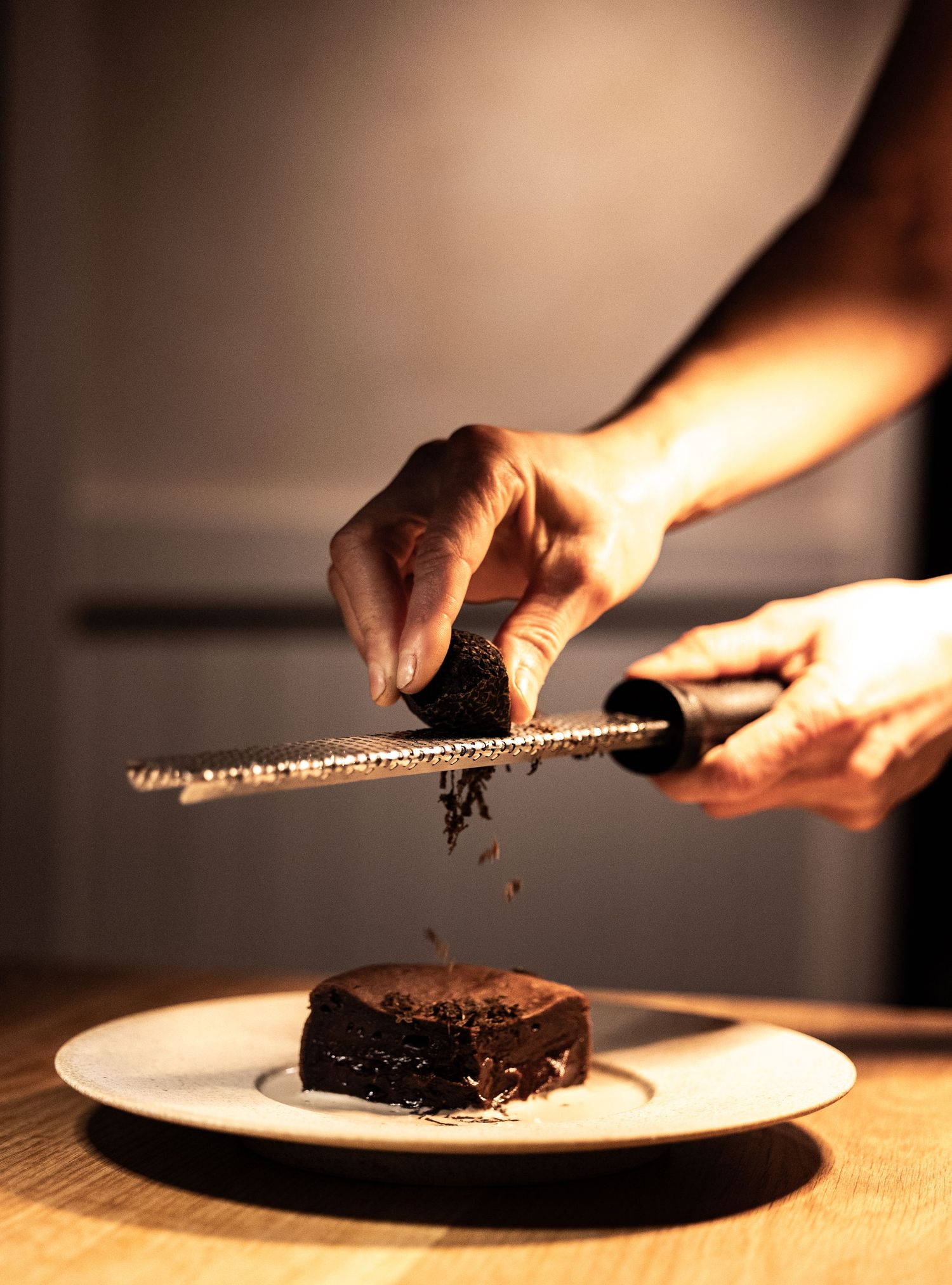 Chef grating dark chocolate truffle over a chocolate dessert on a white plate in a restaurant.