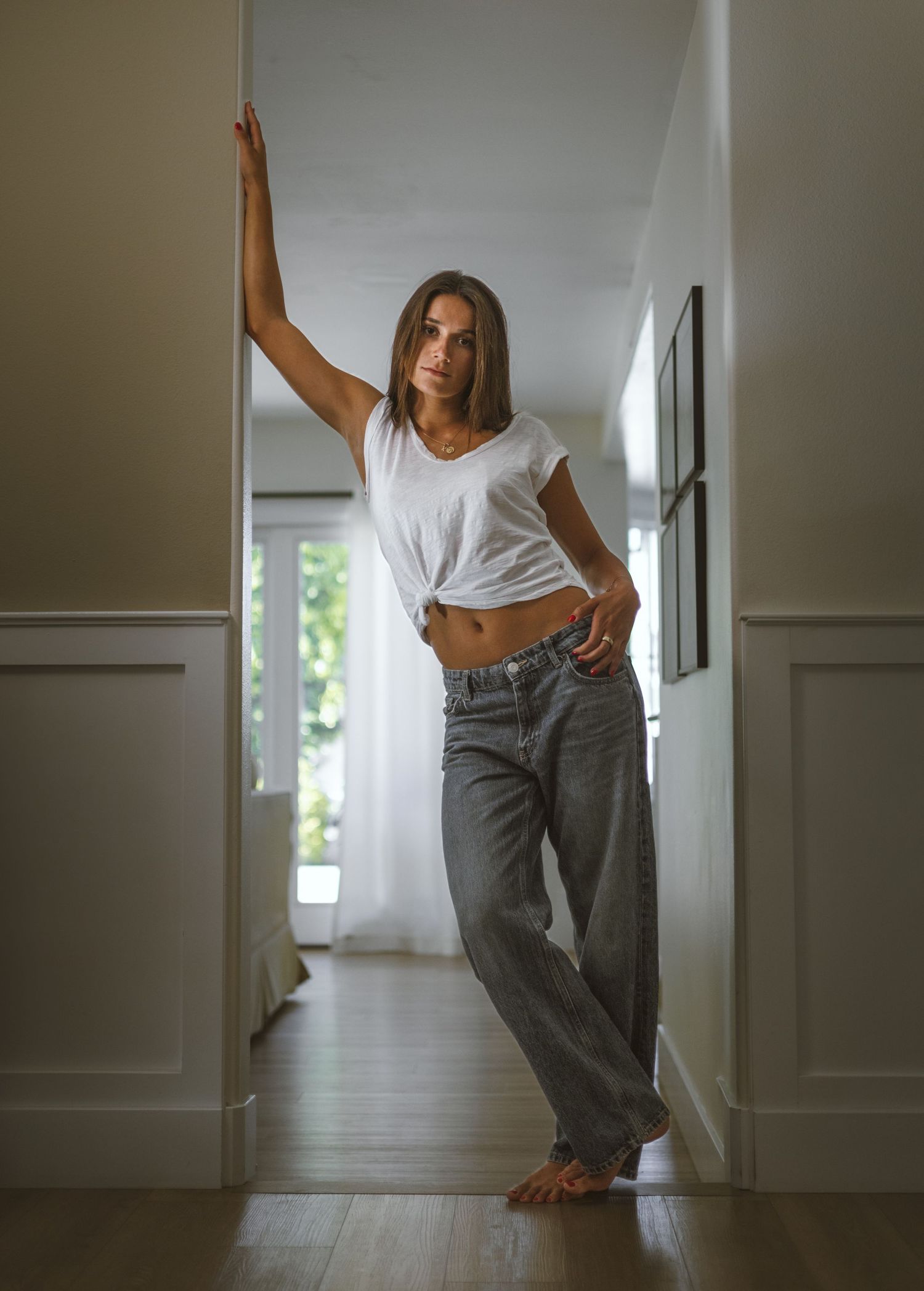 Woman in white crop top and gray jeans posing in a bright hallway doorway.
