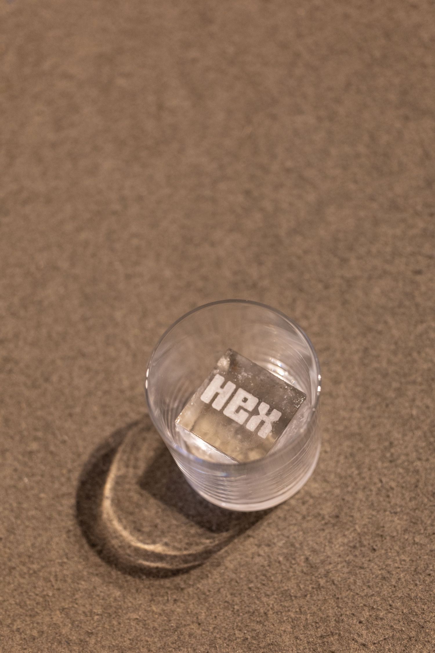 Top view of a glass with ice cube engraved with HEX logo sitting on sandy beach surface.
