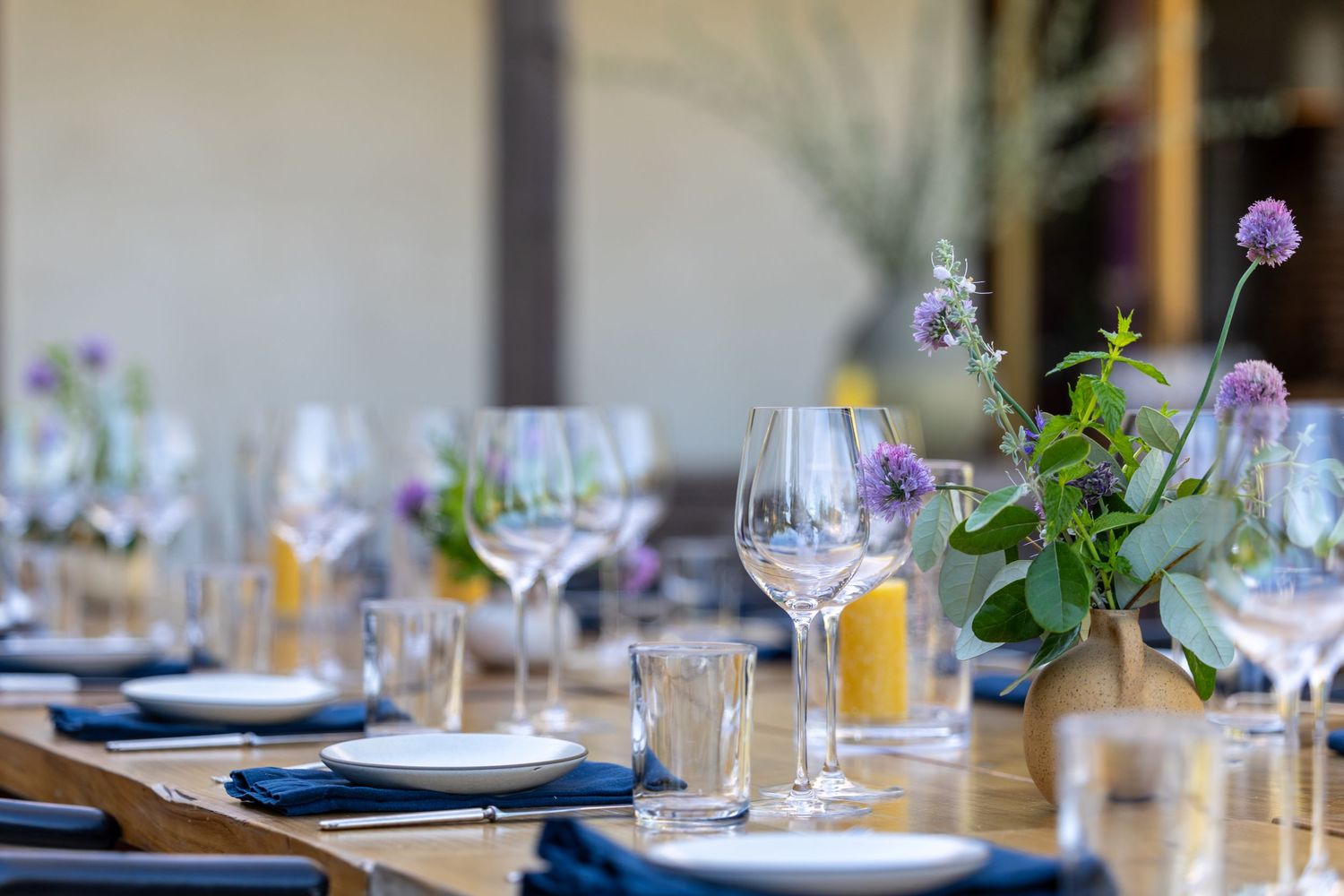 Elegant table setting with wine glasses, blue napkins, and purple floral centerpieces on a wooden dining table.