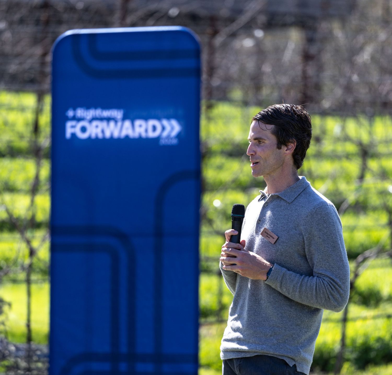 A man speaks into a microphone outdoors at a RightWay Forward event with green fields in the background.