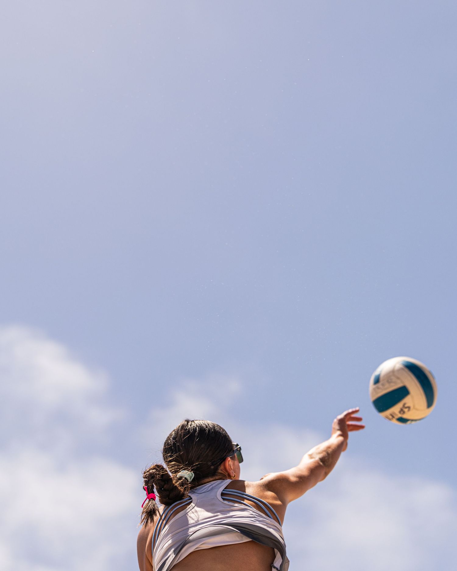 Woman reaching up to spike a volleyball against a bright blue sky during a beach volleyball game.
