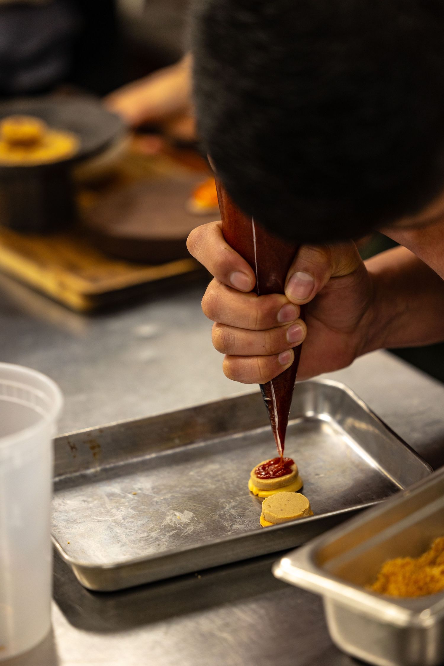 Chef using a piping bag to plate sauce on yellow discs in a professional restaurant kitchen.
