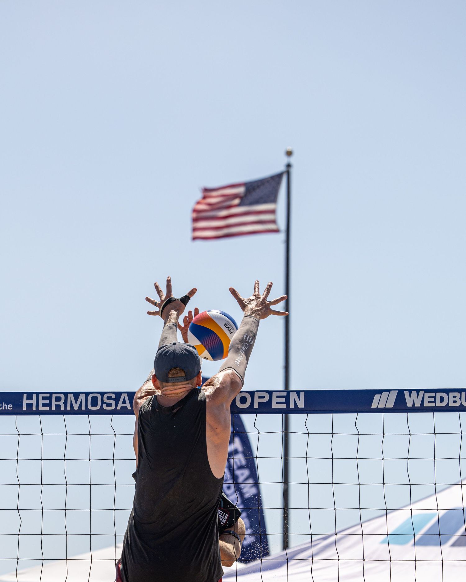 Player blocks volleyball at net during Hermosa Open beach volleyball tournament with US flag waving in background.