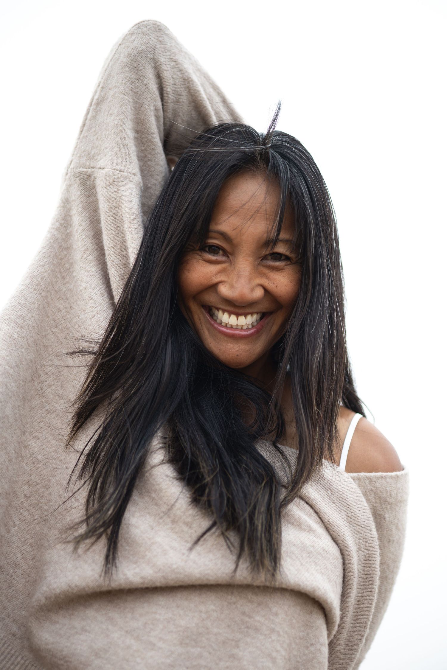 Smiling woman with long black hair wrapped in a cozy beige sweater against a white background.