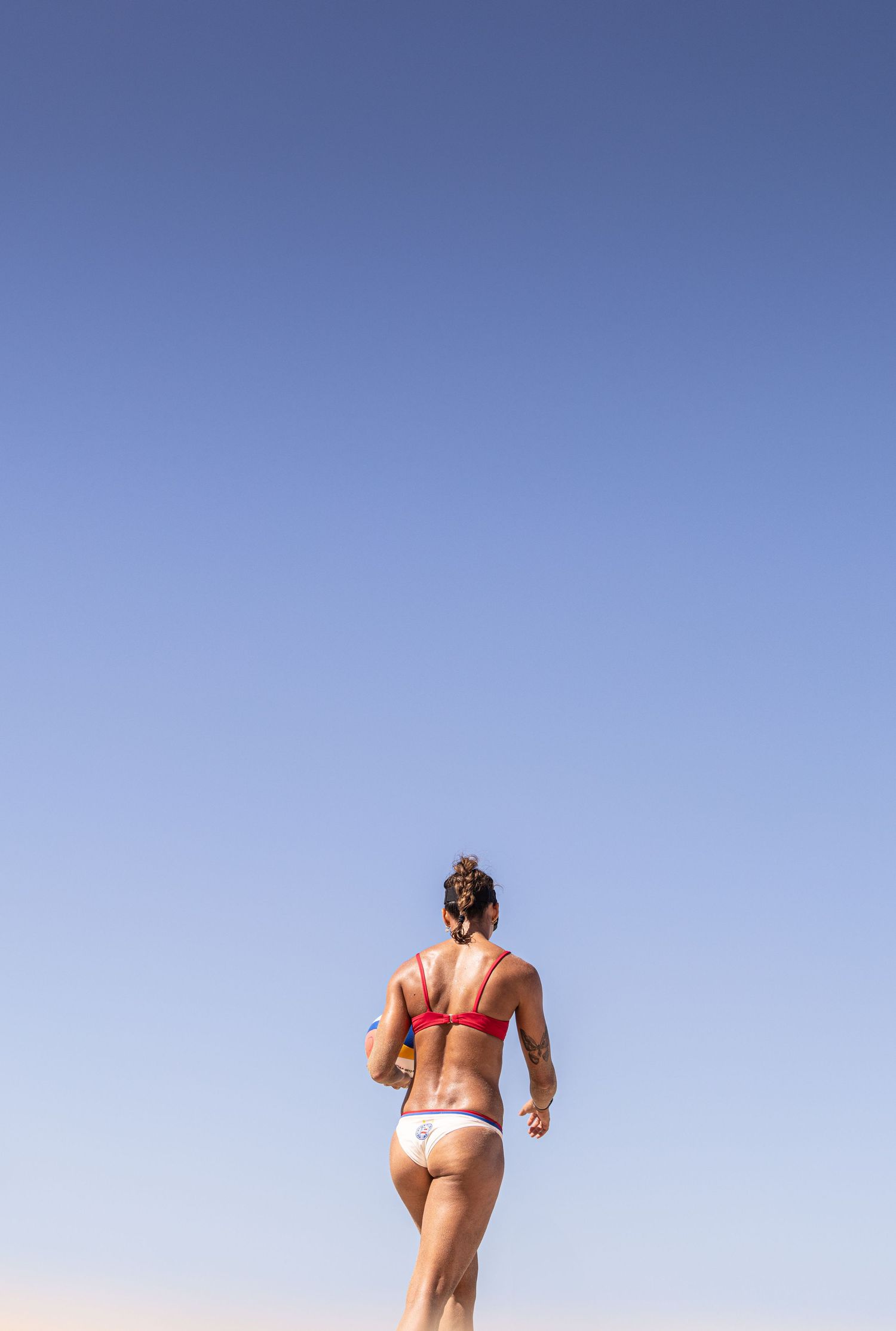 Female beach volleyball player in red bikini viewed from behind against clear blue sky.
