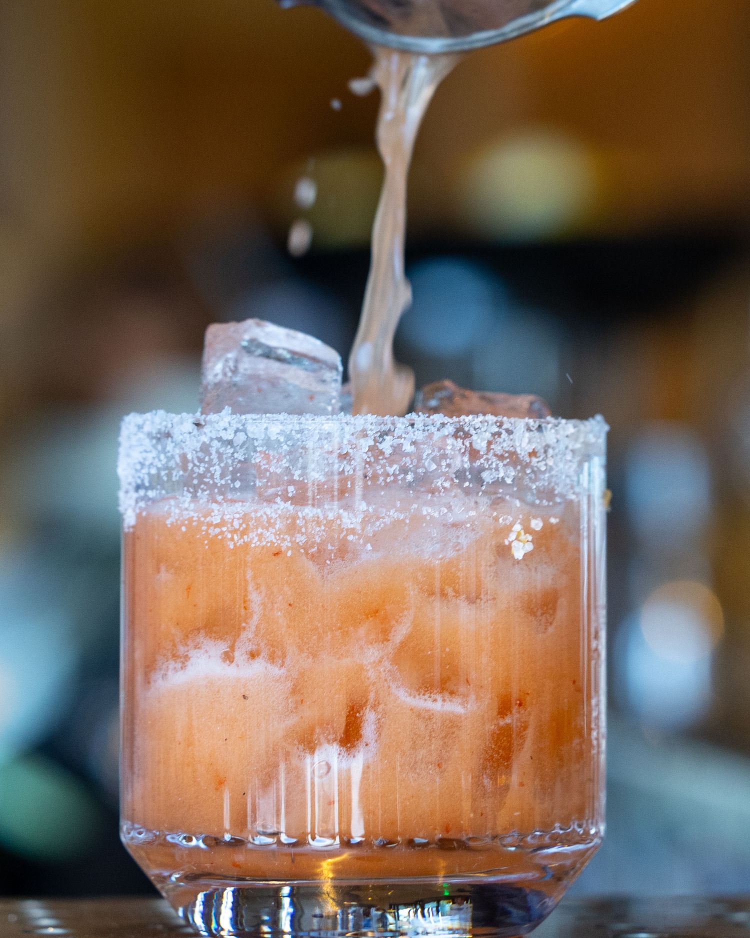 A salted-rim cocktail glass filled with a pink drink and ice being poured at a bar.