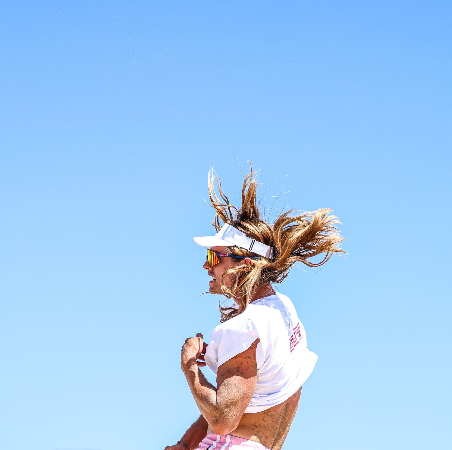 Athletic woman running outdoors wearing white visor and sunglasses against a clear blue sky.