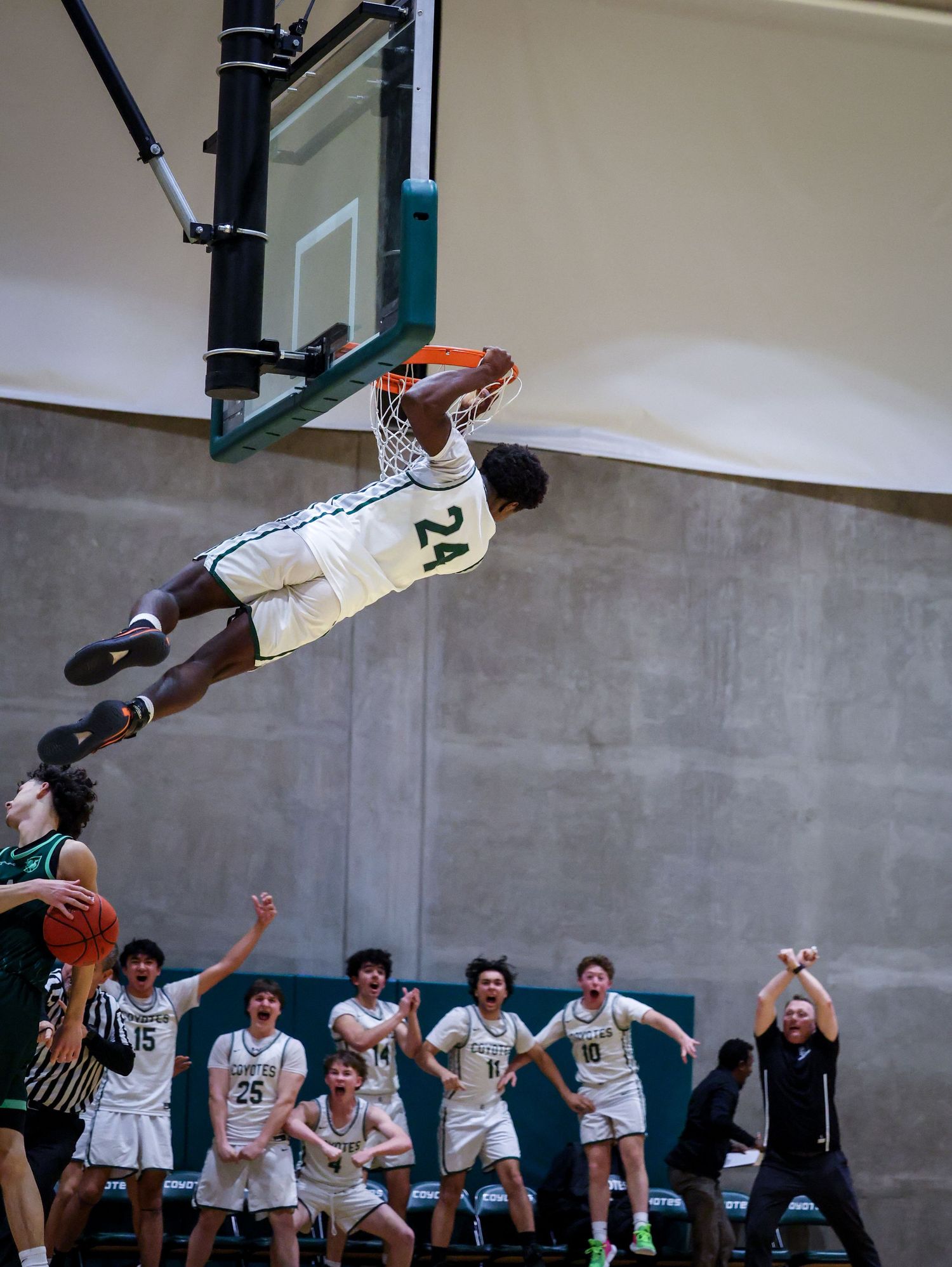 Basketball player #24 dunks dramatically, hanging from rim, as teammates celebrate on the bench below.