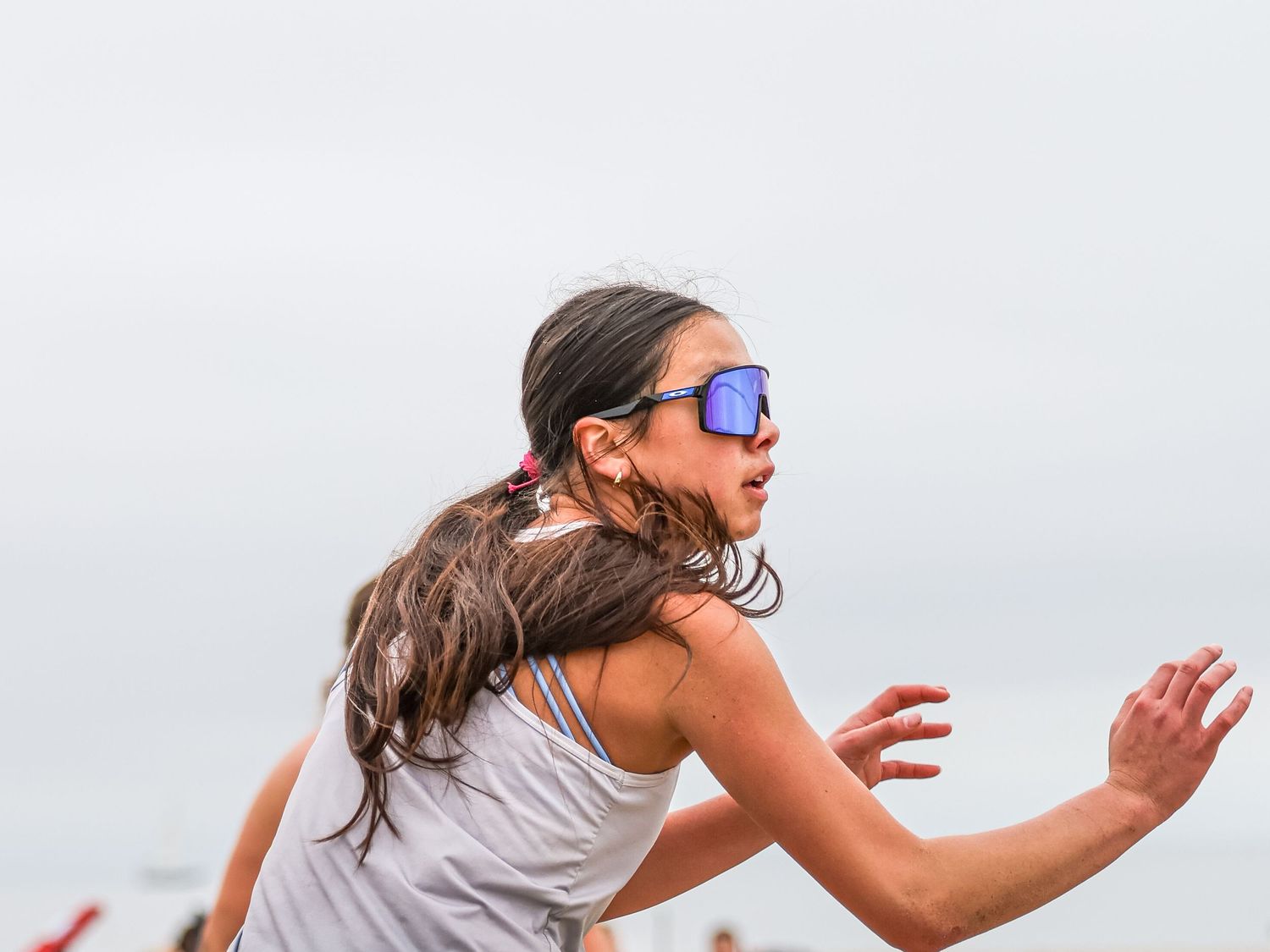A young girl with sunglasses sprints energetically on a beach, her ponytail flying as she runs with arms outstretched.