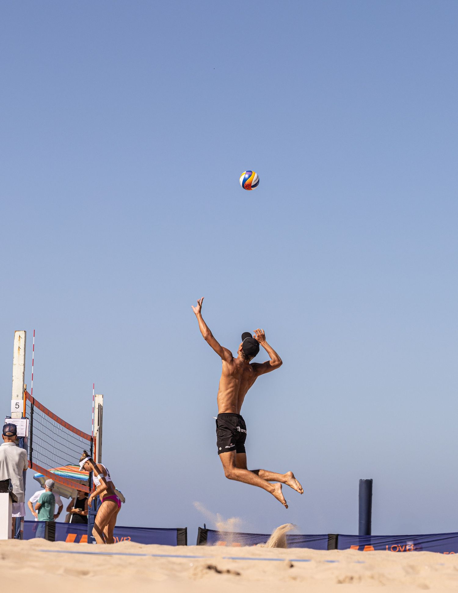 Shirtless male beach volleyball player jumping high to spike ball over net on sandy court under clear blue sky.