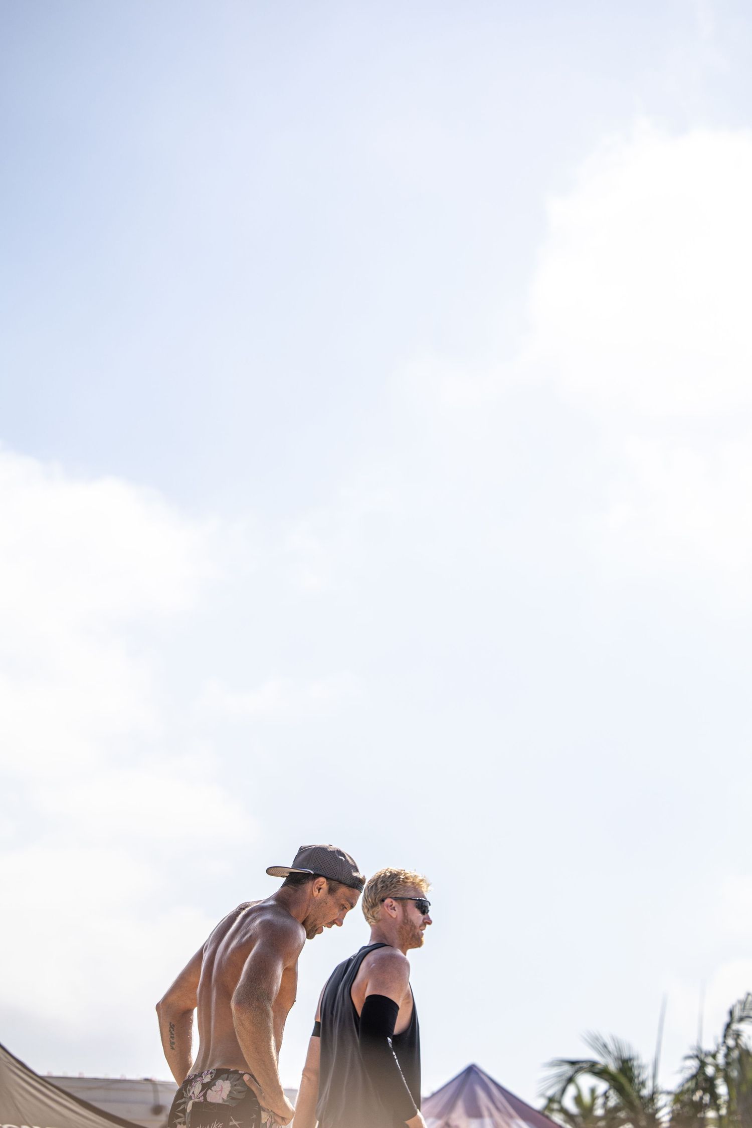Two shirtless men conversing outdoors under a bright sky at a beach festival.