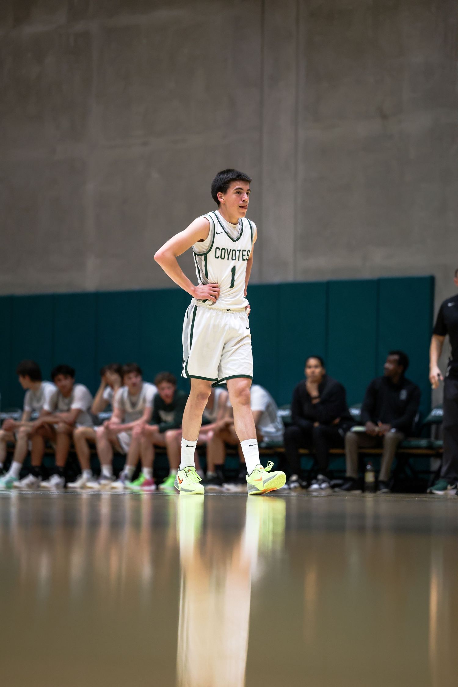 Basketball player in white Coyotes jersey number 1 stands on court during game with teammates on bench behind.