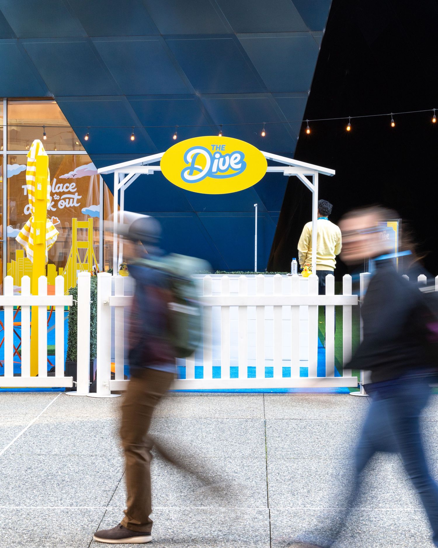 People walk past The Dive branded kiosk with white picket fence and yellow sign at a busy indoor venue.