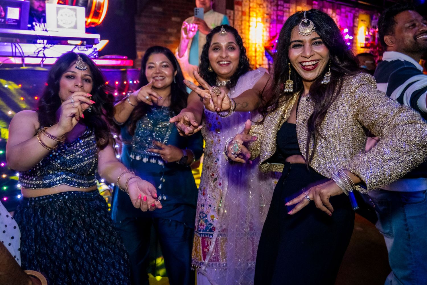 Four women in festive Indian attire dancing joyfully at a colorful party.