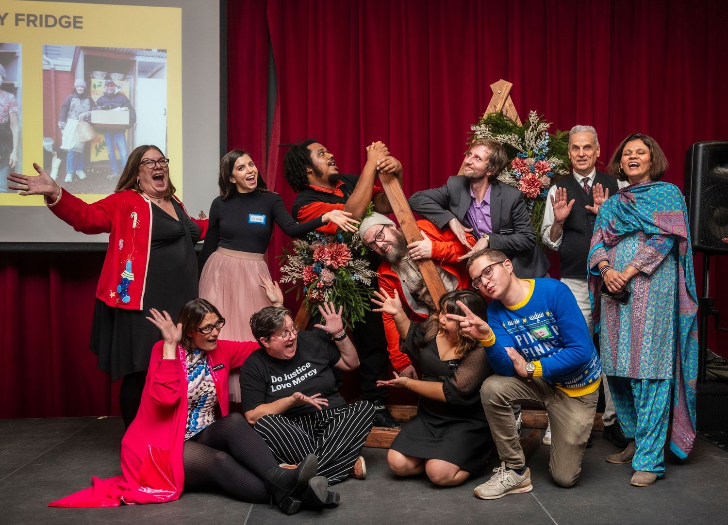 A diverse group of people celebrating together on stage, posing joyfully with raised hands and colorful outfits at an event.