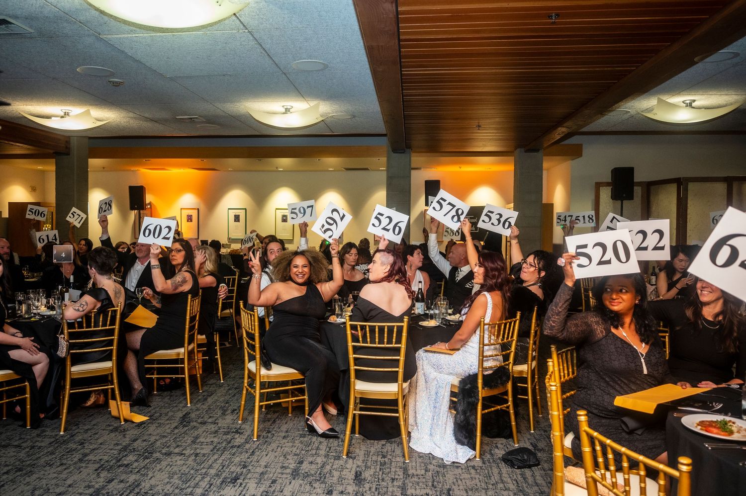 Elegantly dressed women in black raise numbered bidding paddles enthusiastically at a gala auction.