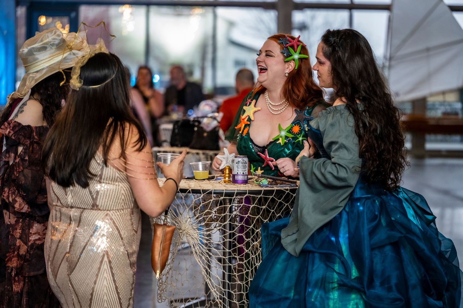 Three women in festive costumes laugh together at a nautical-themed cocktail party.