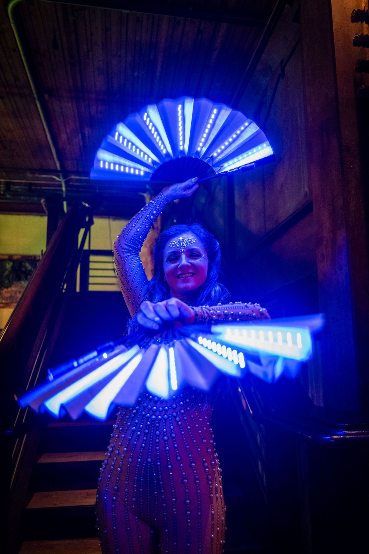 A female performer holds illuminated blue LED fan props while wearing a rhinestone costume in dark lighting.